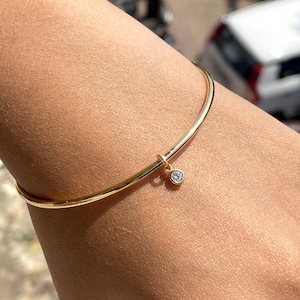 May include: A slender gold-coloured bracelet featuring a tiny, round, clear gemstone pendant. The bracelet is a simple, thin band with a gentle curve, suitable for daily wear. The charm hangs from a small loop.