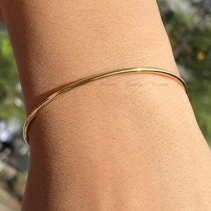 Może przedstawiać: Cienka, polerowana bransoletka typu bangle w kolorze złotym o gładkiej, zaokrąglonej powierzchni. Metalowa obręcz tworzy ciągły okrąg, odbijający światło na swojej zakrzywionej zewnętrznej stronie. Słowo Florence jest powtórzone małym drukiem na metalu.
