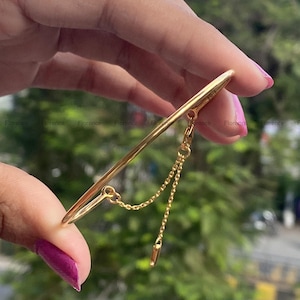 Może przedstawiać: Złota metalowa bransoletka typu bangle z gładkim, zakrzywionym prętem. Do pręta przymocowany jest delikatny, cienki złoty łańcuszek, który zwisa z małą, cylindryczną metalową zawieszką na końcu, tworząc wiszący element ozdobny.