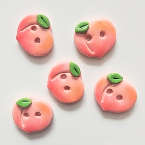 May include: Five peach-shaped buttons with a pink and orange gradient, each featuring a small green leaf and two buttonholes. These decorative buttons are ideal for sewing and craft projects.