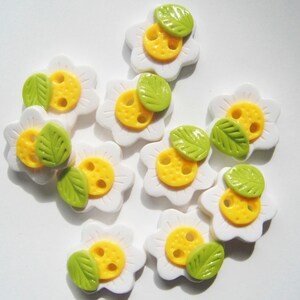 May include: Nine white flower-shaped buttons with yellow centers and green leaves. The buttons have two holes each.