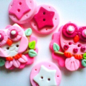 May include: Five pink and white fabric buttons with different designs. The buttons feature an owl, a star, and a circle.