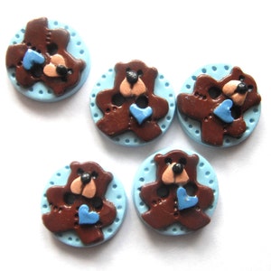 May include: Five brown teddy bear buttons with blue hearts on a light blue background. The buttons are approximately 1 inch in diameter.