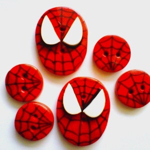 May include: Set of six red and black Spider-Man themed buttons. The buttons feature a web design and a white spider-man mask. Two of the buttons are oval shaped and four are round.