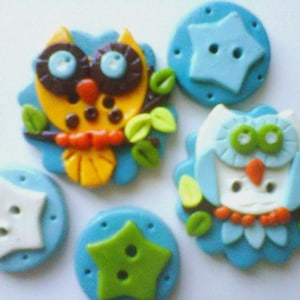May include: A collection of handmade owl and star-shaped buttons. The owl buttons feature yellow, white, and blue owls with orange beaks and green leaves. The star buttons are blue, white, and green. These buttons are ideal for craft projects.