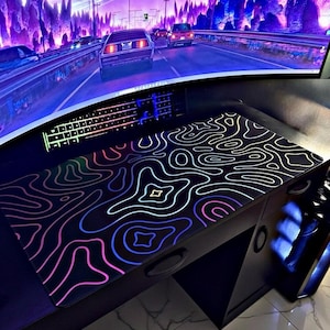 May include: A rectangular desk mat featuring a black surface with a vibrant, multi-colored topographic line pattern in shades of blue, purple, and pink. The mat is positioned on a dark desk surface in front of a computer monitor and keyboard.