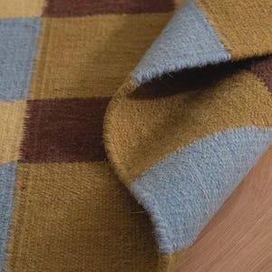 May include: A woven rug featuring a repeating checkered pattern in shades of light blue, mustard yellow, and dark brown. The rug is folded over at one corner, revealing the textured surface and the finished edge of the fabric construction.