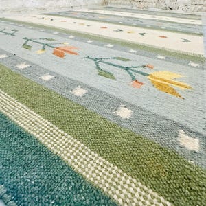 Könnte beinhalten: Ein gewebter Teppich mit horizontalen Streifen in Salbeigrün, Creme und Hellblau. Das Design zeigt wiederkehrende florale Motive in Orange und Gelb, akzentuiert durch kleine weiße quadratische Muster auf der strukturierten Oberfläche.