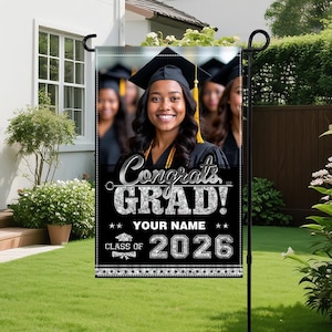 Puede incluir: Una bandera de jardín de graduación negra y plateada con el texto "Congrats GRAD!" y "YOUR NAME" debajo de una foto de una graduada sonriente. La bandera también muestra "CLASS OF 2026". La bandera se muestra en un soporte de metal negro en un jardín.