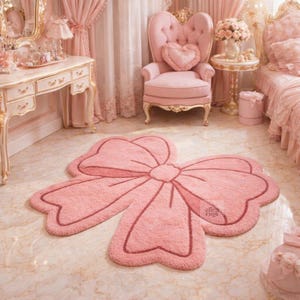 Può includere: Un tappeto rosa a forma di fiocco, con un contorno e un dettaglio centrale rosa più scuro. Il tappeto è su un pavimento di marmo in una stanza con mobili e decorazioni rosa. È un elemento decorativo.
