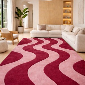 May include: A large, rectangular rug with a wavy pattern in shades of pink and burgundy. The rug is placed on a hardwood floor in a living room setting, with a white sofa and natural light.