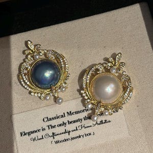 Puede incluir: Dos broches ornamentados con perlas grandes. Uno presenta una perla azul oscuro, el otro una perla blanca, ambos engastados en marcos dorados con detalles brillantes. Los broches forman parte de una caja de joyería de madera.