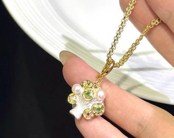 May include: A gold-colored necklace with a pendant shaped like a stylized tree. The pendant features pearls, red and green gemstones, and a white base. The necklace is held by a person.