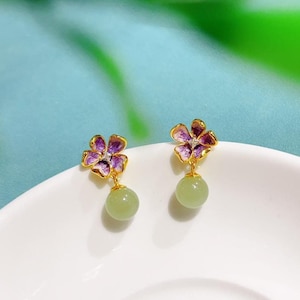 Op de afbeelding: Een paar goudkleurige oorbellen. Elke oorbel heeft een paars en goud bloemmotief met een lichtgroene, ronde kraal die eronder hangt. De oorbellen worden gepresenteerd op een wit oppervlak tegen een blauw-groene achtergrond.