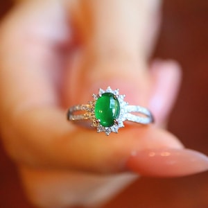 May include: A silver ring featuring a vibrant green oval gemstone, surrounded by a halo of smaller, clear stones. The ring's band splits into two delicate, gem-studded strands on either side of the central stone, creating an elegant design.