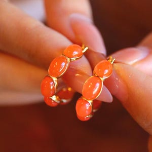 May include: A pair of gold hoop earrings featuring oval-shaped orange gemstones. The earrings are held up close, showcasing the vibrant color and design. The gemstones are set in a gold metal, creating a classic and elegant look.