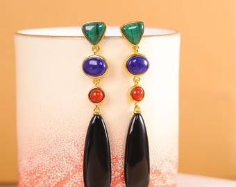 May include: Long dangle earrings with a gold-tone setting. Each earring features a green triangle, a blue oval, a red round stone, and a black teardrop-shaped stone. The earrings are displayed against a white and orange background.