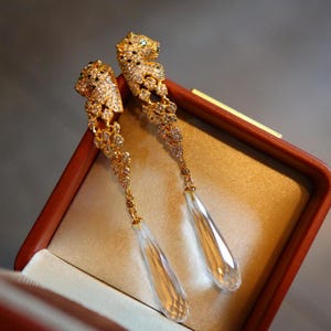 Könnte beinhalten: Ein Paar goldfarbene Ohrhänger mit einem Jaguar-Kopf-Design, besetzt mit kleinen, funkelnden Kristallen. Die Ohrringe haben lange, klare, tropfenförmige Kristallanhänger. Die Ohrringe werden in einer braunen Schmuckschatulle mit goldenem Satinfutter präsentiert.