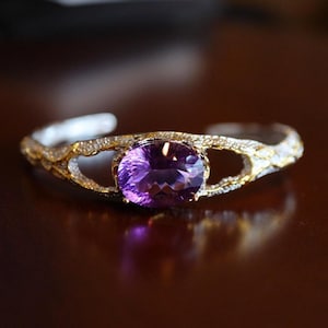 Könnte beinhalten: Ein gold-silbernes Armreif-Armband mit einem großen, oval geschliffenen, lila Amethyst-Edelstein. Das Armband hat ein strukturiertes, offenes Design mit kleinen, funkelnden Akzenten. Der Edelstein ist der Mittelpunkt, gefasst in einer goldenen Lünette.
