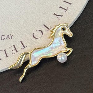 Könnte beinhalten: Goldfarbene Pferdebrosche mit schillerndem Körper und kleiner Perle unten. Das Pferd befindet sich in einer springenden Pose, mit einem schwarzen Edelstein für das Auge. Die Brosche ist vor einem neutralen Hintergrund platziert.