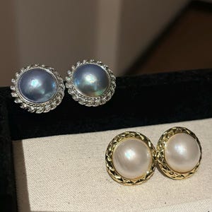 Peut inclure: Deux paires de boucles d'oreilles à clous en perles. La paire supérieure présente des perles grises irisées entourées d'une bordure argentée incrustée. La paire inférieure a des perles blanches avec une bordure dorée texturée. Les boucles d'oreilles sont exposées sur une surface noire.