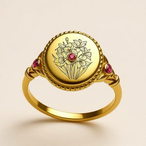 以下が含まれることがあります： 花柄の彫刻と中央の赤い宝石が特徴の、金色のリング。リングの肩には2つの赤い宝石がセットされています。リングは磨かれた仕上げで、クラシックなデザインです。