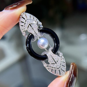 Könnte beinhalten: Eine silberfarbene Brosche mit einer zentralen weißen Perle, die in einem schwarzen kreisförmigen Ring hängt. Die Brosche ist mit komplizierten silbernen Filigranmustern verziert, die an beiden Enden mit zahlreichen kleinen, klaren Steinen besetzt sind.