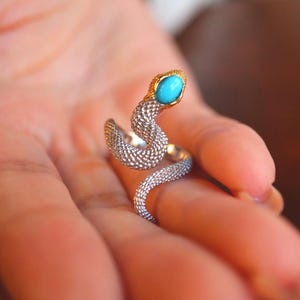 Op de afbeelding: Zilveren slangenring met een turquoise edelsteen kop en goudkleurige accenten. De ring is ontworpen om de vinger te omsluiten, wat een uniek en opvallend sieraad creëert.