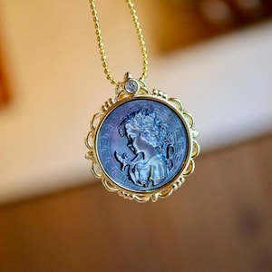 Peut inclure: Un collier pendentif doré avec un camée circulaire bleu-gris irisé représentant le profil d'un enfant. Le camée est serti dans un cadre doré décoratif avec un petit diamant. La chaîne du collier est également dorée.