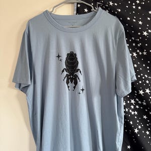May include: A light blue t-shirt with a black insect graphic and star accents. The shirt is hanging on a metal hanger. The background includes a black and white star-patterned fabric.