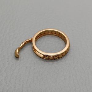 May include: A gold-colored ring with a unique design. The ring features a circular band with small, evenly spaced decorative elements. A curved, textured extension adds an artistic flair to the piece. The ring is set against a gray background.