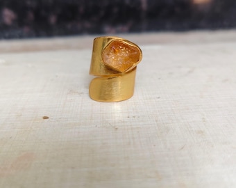 May include: A gold-toned ring featuring a wide, brushed metal band with an open-ended wrap design. The top of the ring is set with a single, irregular, translucent amber-colored gemstone with a rough, textured surface.