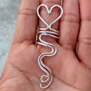 Puede incluir: Un anillo plateado con un diseño en forma de corazón en la parte superior y un diseño ondulado y fluido. El diseño del anillo se extiende por el dedo, creando un aspecto único y artístico. El anillo se muestra en una mano.
