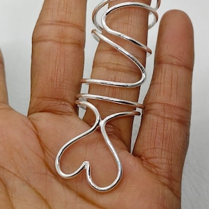 May include: A silver-colored ring with a unique design. The ring features a heart-shaped base and a swirling, elongated upper portion that wraps around the finger. The ring is made of a shiny, metallic material.