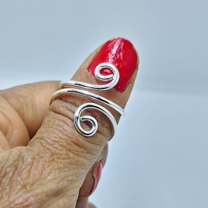 May include: A silver-colored, adjustable ring with a spiral design. The ring wraps around a finger, featuring two decorative swirls. The background is a light gray, and the fingernail is painted red.