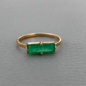 May include: A gold ring featuring a rectangular, emerald-cut green gemstone. The ring has a thin, gold band and small gold accents on either side of the emerald. The ring is set against a neutral background.