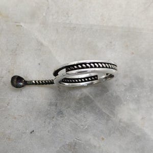 May include: A silver ring with a rope-like design around the band. The ring has a small, decorative spoon attached to it. The spoon has a small, round bowl and a twisted handle. The ring is on a light-colored surface.