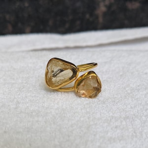 Peut inclure: Une bague dorée ornée de deux pierres jaunes translucides aux formes irrégulières, serties dans des montures en métal. Le design présente un anneau fin et ouvert reliant les deux gemmes brutes, offrant un aspect superposé et asymétrique.
