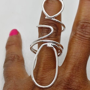 May include: A silver ring with an abstract design. The ring features a large oval shape and a smaller circular shape connected by curved lines. The ring is worn on a finger with pink nail polish.
