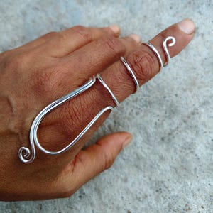 Könnte beinhalten: Ein silberfarbener Fingerring mit einem einzigartigen, offenen Design. Der Ring hat ein Spiraldetail an der Fingerspitze und eine gebogene, längliche Form, die sich den Finger hinunter erstreckt. Der Schmuck wird auf einer Hand vor neutralem Hintergrund gezeigt.