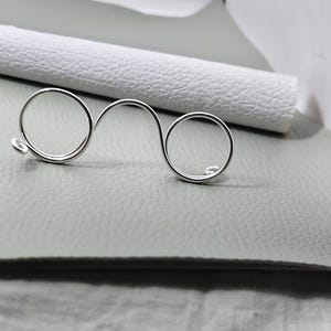 May include: A silver ring featuring a double-circle design, joined by a curved bar. The ring sits on a light grey surface, with a rolled white textured material behind it. The ring's design is minimalist and elegant, suitable for everyday wear.