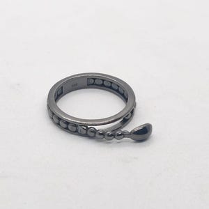 以下が含まれることがあります： ユニークなデザインのダークグレーのリング。リングは、小さく均等に配置されたビーズと、ぶら下がっている涙の形をしたチャームが付いたバンドが特徴です。リングは白い背景に配置されています。
