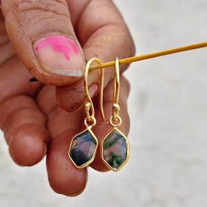 May include: Gold-colored dangle earrings with hexagonal, gemstone pendants. The gemstones have a mix of dark green and maroon colors. The earrings are held up by a wooden stick.