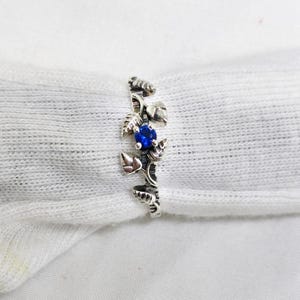 Può includere: Un anello d'argento con un design floreale, con foglie e una gemma blu ovale centrale. L'anello è posto su uno sfondo bianco, evidenziando l'intricato lavoro del metallo e il colore vibrante della pietra.