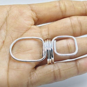 May include: A silver ring with two rectangular open shapes connected by a series of smaller bands. The ring is displayed on a hand, showcasing its design and craftsmanship. The ring's polished surface reflects light, highlighting its metallic sheen.