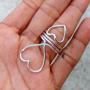 May include: A silver-colored ring with a heart-shaped design. The ring features two heart shapes connected by a series of bands. The ring is worn on a finger.