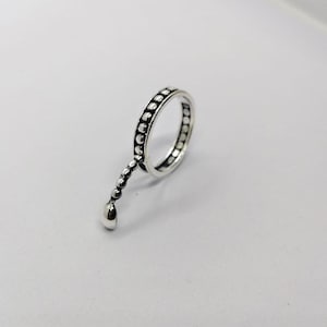 May include: A silver ring with a beaded design around the band and a dangling teardrop-shaped charm. The ring is set against a white background, highlighting its intricate details and polished finish.