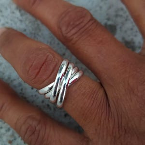May include: A silver ring with a crisscross design. The ring features four bands that intersect, creating a unique and modern look. The polished metal reflects light, highlighting the ring's smooth, rounded surfaces. The ring is worn on a finger.