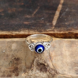 Puede incluir: Anillo plateado con un diseño de ojo azul y pupila negra. El anillo tiene detalles laterales decorativos y está sobre una superficie de madera rústica. Una joya con un diseño único y simbólico.