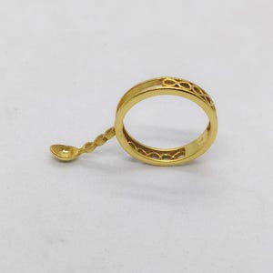 May include: A gold-colored ring with a decorative band and a small spoon attached. The ring band features an openwork design. The spoon is connected to the ring with a twisted stem. The ring is set against a white background.